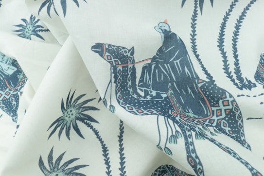 A fabric with a printed pattern featuring a person dressed in traditional clothing riding a camel. The design includes palm trees and decorative motifs in an artistic style, using a limited color palette.