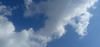 White clouds scattered across a clear blue sky, with sunlight illuminating the edges of the clouds, creating soft shadows and highlights.