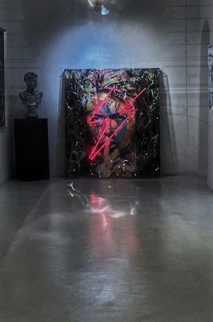 A dimly lit art gallery room featuring a colorful, expressive painting of a face with vibrant neon red lightning bolt streaks across it. A spotlight illuminates the painting, adding a glowing effect. To the left, a sculpture bust is visible, adding to the artistic ambiance of the room.