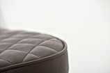 A close-up of a quilted, leather-like surface with a soft, cushioned texture. The stitching forms a pattern of square shapes, and the material appears to be dark gray with a smooth finish. The background is blurred, creating a minimalist and focused composition.