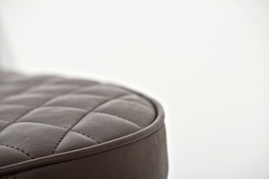 A close-up of a quilted, leather-like surface with a soft, cushioned texture. The stitching forms a pattern of square shapes, and the material appears to be dark gray with a smooth finish. The background is blurred, creating a minimalist and focused composition.