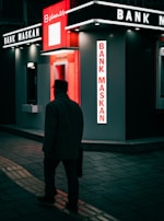 A person walks past a modern, illuminated bank kiosk in an urban setting. The signage is prominently displayed with the name 'Bank Maskan' in bright red and white colors. The atmosphere is dark with artificial lighting creating a contrast against the surroundings.