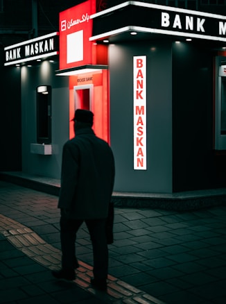 A person walks past a modern, illuminated bank kiosk in an urban setting. The signage is prominently displayed with the name 'Bank Maskan' in bright red and white colors. The atmosphere is dark with artificial lighting creating a contrast against the surroundings.
