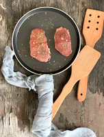 A rustic cast iron skillet sizzling with golden-brown seared steaks on a wooden table.