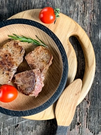 A wooden table with two round wooden cutting boards. On the smaller board, there are three pieces of grilled meat garnished with a sprig of rosemary. Two red tomatoes are placed on the board for decoration.
