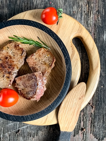 A wooden table with two round wooden cutting boards. On the smaller board, there are three pieces of grilled meat garnished with a sprig of rosemary. Two red tomatoes are placed on the board for decoration.