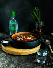 A dimly lit table with eerie purple lighting showcasing a steaming bowl of dark soup surrounded by shadowy decor.