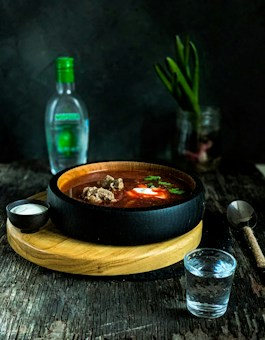 A dark wooden table holds a wooden serving board with a black bowl filled with a rich, dark soup. Pieces of meat, a dollop of sour cream, and herbs float in the soup. To the side, a small black bowl contains more sour cream. A clear glass of water sits in front of the bowl. In the background, a glass bottle with a green cap and a jar with green stalks are visible. The setting is dimly lit, creating a cozy and inviting atmosphere.