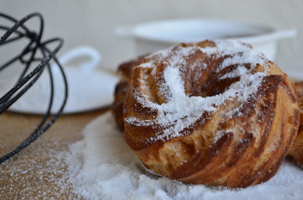 Two baked pastries covered in powdered sugar rest on a wooden surface. A black metal whisk lies nearby, and a white ceramic dish is visible in the background, slightly out of focus.
