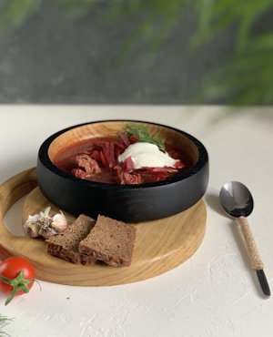 A wooden bowl filled with a hearty, red soup garnished with a dollop of white cream and fresh dill rests on a circular wooden board. Next to the bowl are two slices of brown bread, a clove of garlic, and a ripe red tomato with a vine. A spoon with a wrapped handle lies beside the board on a textured white surface. The background features a green, blurred foliage.