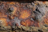 A close-up view of rusted and corroded metal displaying rough textures and patterns. The surface features various shades of orange, brown, and black with noticeable patches of flaking rust, giving it a weathered appearance.