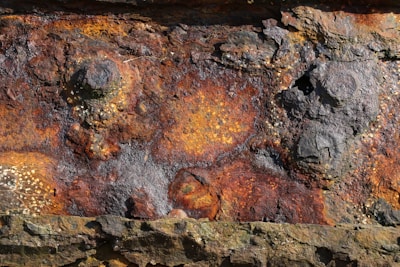 A close-up view of rusted and corroded metal displaying rough textures and patterns. The surface features various shades of orange, brown, and black with noticeable patches of flaking rust, giving it a weathered appearance.