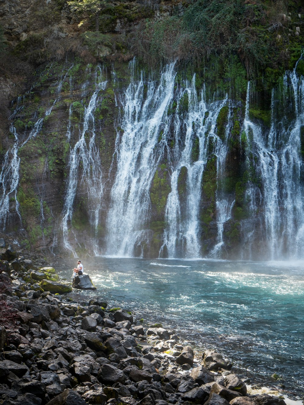 woman in yellow shirt sitting on rock near waterfalls during daytime