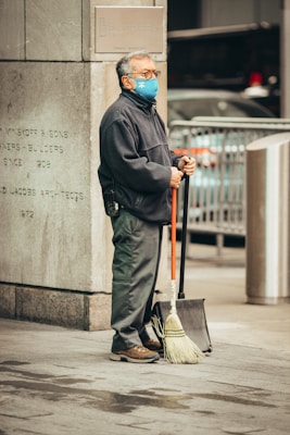 A person wearing a blue face mask, glasses, and a dark jacket stands on a city sidewalk. They are holding a broom and a dustpan. There is a stone wall with some engraved text and a metal sign in the background. A vehicle is partially visible behind a metal barricade.