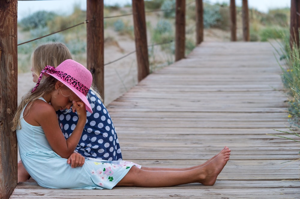 girl in blue and white polka dot shirt and pink hat sitting on wooden bridge