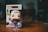 A Funko Pop figurine stands on a wooden surface. The figure has a superhero costume with a blue suit, red gloves, and boots. The character's blonde hair is stylized, and it is displayed in front of its original packaging box, which is slightly blurred in the background.