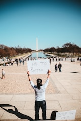A person is holding a sign that reads 'NO TO RACISM' in front of the Washington Monument. The scene is set in an urban park with a reflecting pool, surrounded by trees and numerous people walking or interacting with each other.