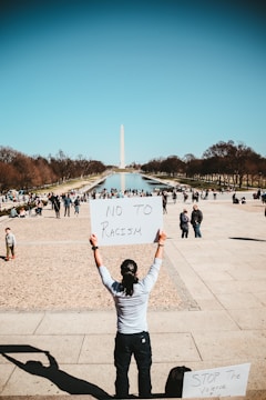 A person is holding a sign that reads 'NO TO RACISM' in front of the Washington Monument. The scene is set in an urban park with a reflecting pool, surrounded by trees and numerous people walking or interacting with each other.