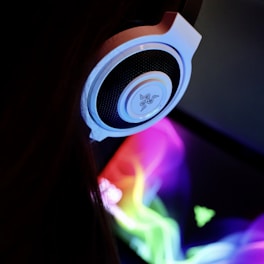 A close-up view of a white headset with a black speaker cover against a backdrop of a colorful and vibrant display. The display features swirling patterns of multicolored light, creating a dynamic and engaging visual effect.