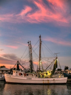 A fishing boat named 'Charlee Girl' is docked with visible nets and rigging. The sky is painted with vibrant pink and blue hues as the sun sets.