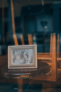 A framed sign containing the words 'Black Lives Matter' and a raised fist is placed on a round wooden table in front of upside down chairs, likely suggesting a closed cafe or restaurant.