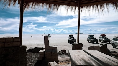 A serene landscape featuring a vast salt flat under a bright blue sky with scattered clouds. Several off-road vehicles with luggage atop are parked on the salt surface. The foreground is shaded by a thatched roof structure with rustic stone furniture, providing a stark contrast to the sunlit expanse beyond.