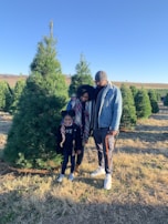 A family of three stands together in a field of pine trees. They appear to be enjoying an outing, with the child smiling brightly while holding hands with the adults. The man and woman are dressed warmly and are looking affectionately at each other and the child. The background features rows of lush green pine trees under a clear blue sky.