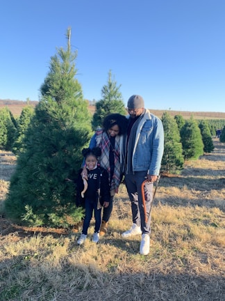 A family of three stands together in a field of pine trees. They appear to be enjoying an outing, with the child smiling brightly while holding hands with the adults. The man and woman are dressed warmly and are looking affectionately at each other and the child. The background features rows of lush green pine trees under a clear blue sky.