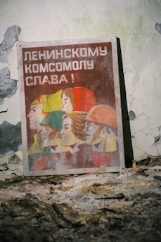 An old, worn poster leaning against a peeling wall, featuring stylized illustrations of people in profile with vibrant colors like red, yellow, and green. Russian text is prominently displayed above the figures.