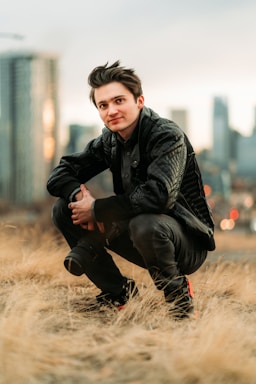 A person with short dark hair squats on dry grass, holding a camera. The individual wears a black jacket and pants against an urban skyline backdrop with blurred buildings.