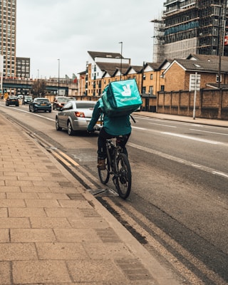 A cyclist wearing casual clothing and a large turquoise delivery backpack is riding along a city street. There are several vehicles on the road and modern apartment buildings in the background. The atmosphere is urban and slightly overcast.