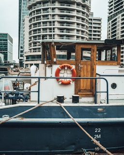 A medium-sized boat is moored at a dock with a wooden cabin and a life preserver labeled 'Anita' on its side. The boat features a rustic wooden door and is painted in shades of blue and white. In the background, modern high-rise buildings with unique architecture are visible, indicating an urban waterfront setting. Ropes are tied to the boat, securing it in place.