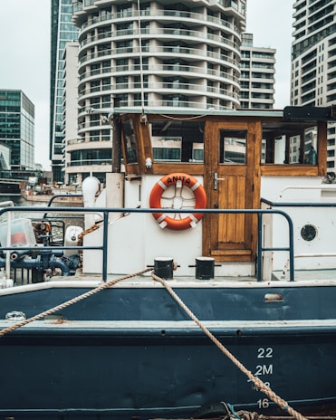 A medium-sized boat is moored at a dock with a wooden cabin and a life preserver labeled 'Anita' on its side. The boat features a rustic wooden door and is painted in shades of blue and white. In the background, modern high-rise buildings with unique architecture are visible, indicating an urban waterfront setting. Ropes are tied to the boat, securing it in place.