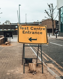 A yellow sign indicating the entrance to a test center is prominently displayed on a sidewalk. The sign features bold black text with an arrow pointing to the left. Sandbags are used to weigh down the metal frame of the sign. The surrounding environment includes a paved sidewalk, a road, bare trees, and urban buildings.