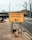 A yellow sign indicating the entrance to a test center is prominently displayed on a sidewalk. The sign features bold black text with an arrow pointing to the left. Sandbags are used to weigh down the metal frame of the sign. The surrounding environment includes a paved sidewalk, a road, bare trees, and urban buildings.