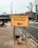 A yellow sign indicating the entrance to a test center is prominently displayed on a sidewalk. The sign features bold black text with an arrow pointing to the left. Sandbags are used to weigh down the metal frame of the sign. The surrounding environment includes a paved sidewalk, a road, bare trees, and urban buildings.