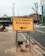 A yellow sign indicating the entrance to a test center is prominently displayed on a sidewalk. The sign features bold black text with an arrow pointing to the left. Sandbags are used to weigh down the metal frame of the sign. The surrounding environment includes a paved sidewalk, a road, bare trees, and urban buildings.