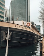 A large boat with the name 'Le Sorelle' moored in an urban waterway. The angle captures the boat from the front and side, with a prominent anchor chain in view. Skyscrapers with reflective glass exteriors tower in the background, contributing to a metropolitan setting. The presence of life preservers indicates a focus on safety.