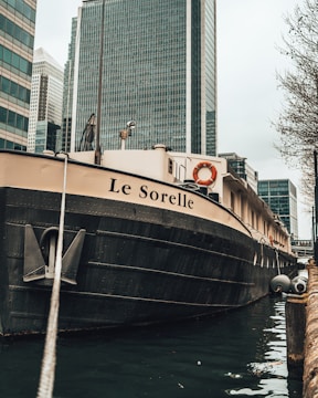 A large boat with the name 'Le Sorelle' moored in an urban waterway. The angle captures the boat from the front and side, with a prominent anchor chain in view. Skyscrapers with reflective glass exteriors tower in the background, contributing to a metropolitan setting. The presence of life preservers indicates a focus on safety.