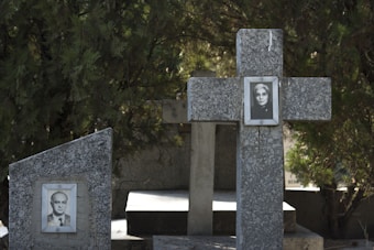 Several stone grave markers in a cemetery, with images of two individuals displayed on them. One is a cross-shaped marker with a woman's photo, and another adjacent marker features a man's photo. The background includes dense, green foliage.