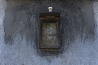 A weathered, rectangular frame encloses an old, monochrome photograph showing a person with a faint, visible face. The frame is mounted on a textured, gray wall, and has a small white knob at the top.