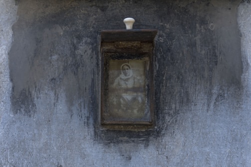 A weathered, rectangular frame encloses an old, monochrome photograph showing a person with a faint, visible face. The frame is mounted on a textured, gray wall, and has a small white knob at the top.
