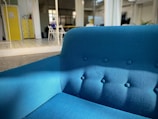 A bright blue upholstered sofa sits prominently in a modern, open space with natural lighting. The room features a yellow cabinet, some chairs, and plants in the background. The setting is contemporary and minimalist.