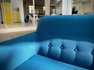 A bright blue upholstered sofa sits prominently in a modern, open space with natural lighting. The room features a yellow cabinet, some chairs, and plants in the background. The setting is contemporary and minimalist.