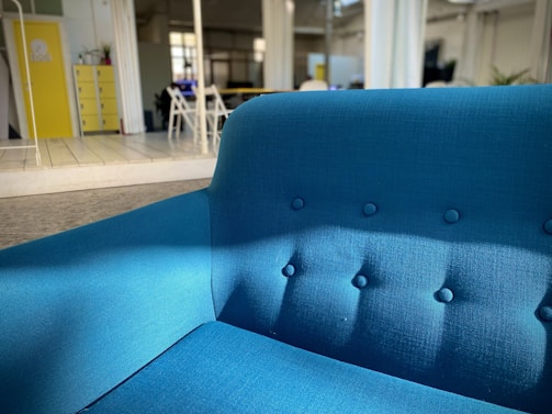 A bright blue upholstered sofa sits prominently in a modern, open space with natural lighting. The room features a yellow cabinet, some chairs, and plants in the background. The setting is contemporary and minimalist.