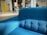 A bright blue upholstered sofa sits prominently in a modern, open space with natural lighting. The room features a yellow cabinet, some chairs, and plants in the background. The setting is contemporary and minimalist.