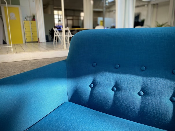 A bright blue upholstered sofa sits prominently in a modern, open space with natural lighting. The room features a yellow cabinet, some chairs, and plants in the background. The setting is contemporary and minimalist.