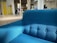 A bright blue upholstered sofa sits prominently in a modern, open space with natural lighting. The room features a yellow cabinet, some chairs, and plants in the background. The setting is contemporary and minimalist.