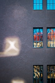 A building façade with textured dark gray paint and several rectangular windows, reflecting vibrant blue and orange hues from the surroundings. The surface is illuminated by patches of soft, diffused light creating abstract shapes.