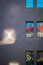 A building fa&ccedil;ade with textured dark gray paint and several rectangular windows, reflecting vibrant blue and orange hues from the surroundings. The surface is illuminated by patches of soft, diffused light creating abstract shapes.